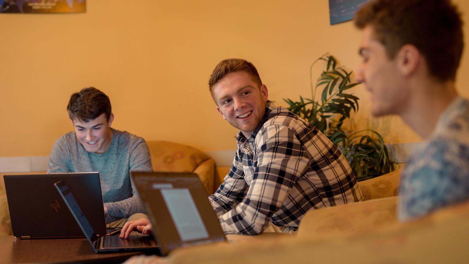 三个学生坐在舒适的椅子上，微笑着在笔记本电脑上工作.